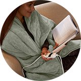 A woman reading a book sitting on a sofa with Silentnight Luxury Heated Throw draped around her. 