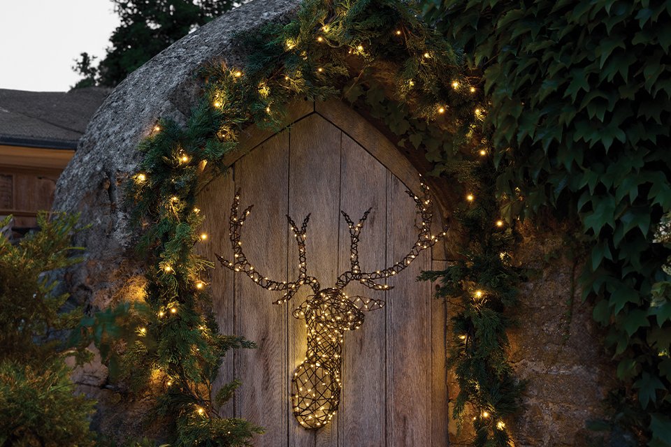 Light up stag head hanging on an outside gate surrounded by a garland with lights 