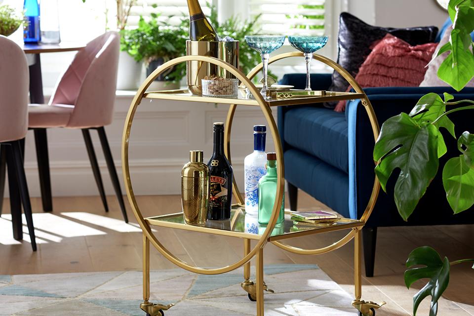 A Habitat drinks trolley in brass finish with a cocktail shaker, ice bucket, champagne and glassware on it.