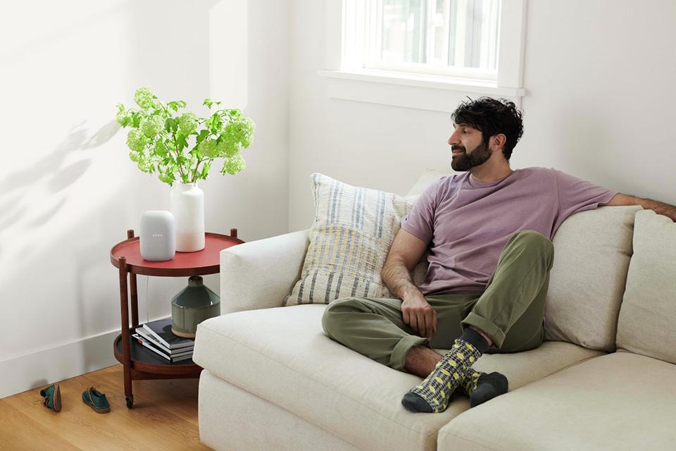 A man sitting on a white sofa looking at a grey Google nest smart speaker kept on the side table.