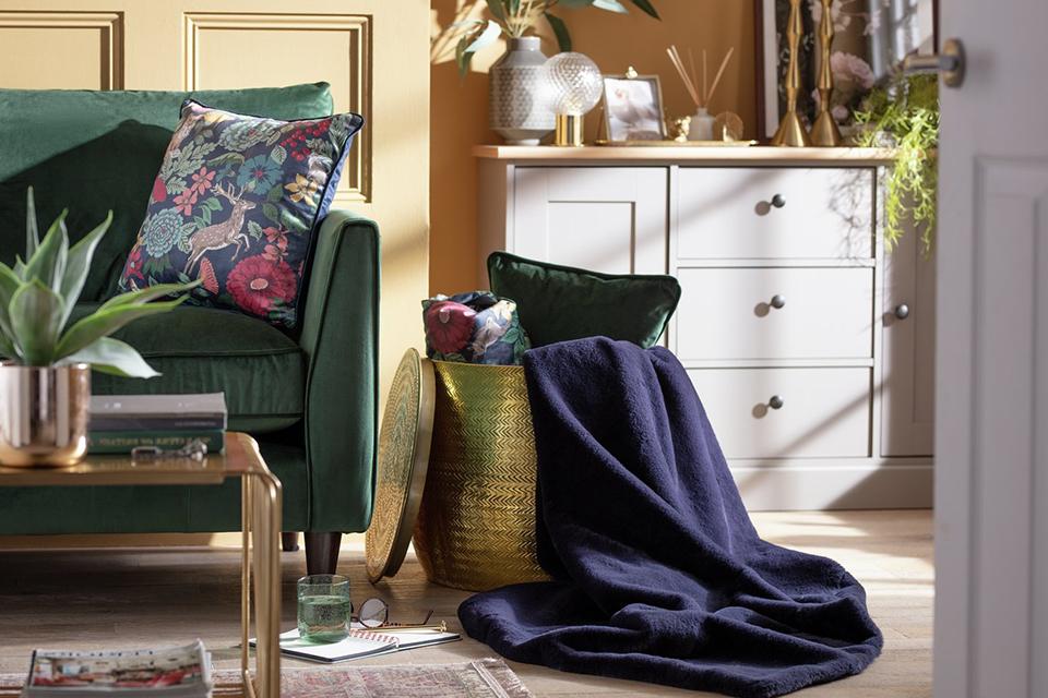 Image of a basket of cushions and blankets next to a green velvet sofa.