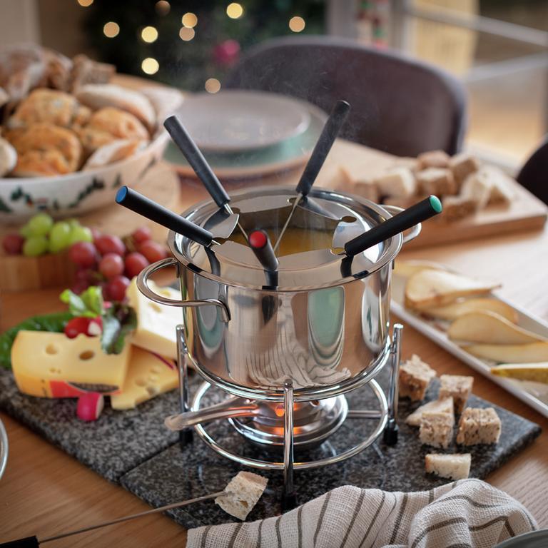 A cheese fondue kept at the centre of a dining table with 5 fondue forks in it.