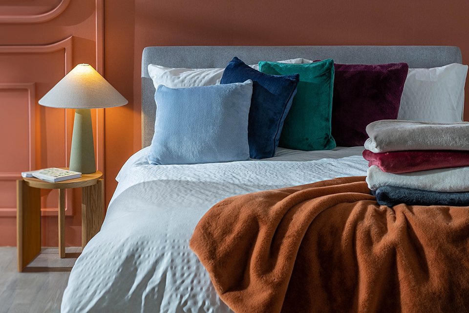 A cosy bedroom with a table lamp on the bedside, throw pillows, cushions and a throw on the bed.