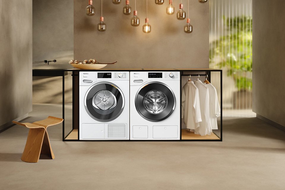 A white Miele washing machine and tumble dryer placed next to each other in an earthy toned room. 