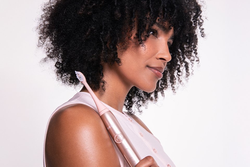 A woman holding a pink Ordo SonicPlus toothbrush against pale pink background.
