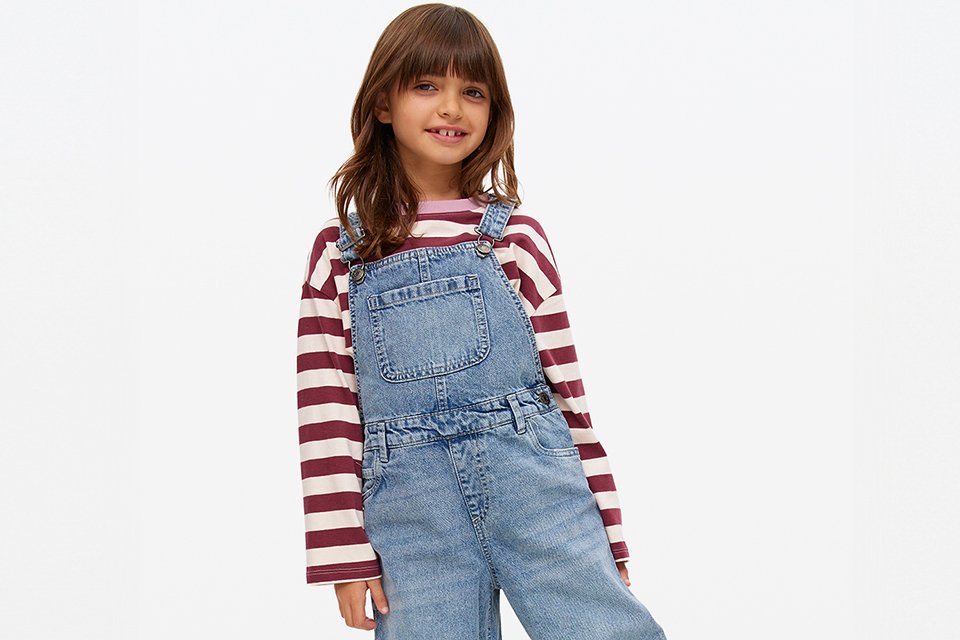 A 7 year old girl in a red t-shirt and mid washed barrel leg denim dungarees.