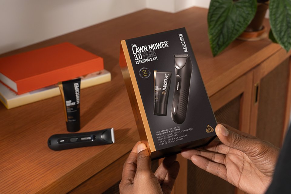 Shot of a person holding Manscaped The Lawn Mover 3.0 Plus box with the product resting on a table.
