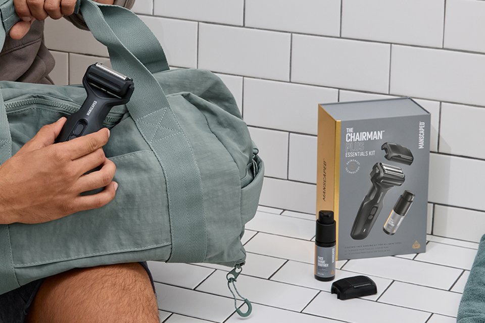 A man holding a duffel bag in one hand and Manscaped Chairman Plus Essentials kit scattered across the bench.