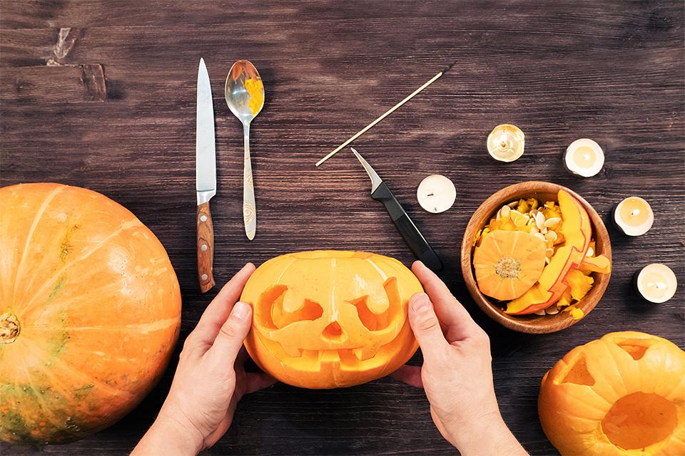 Three pumpkins, with one pumpkin already carved, another ready to be carved and the middle pumpkin being held. Pumpkin parts in a bowl to the right, with utensils surrounding it.