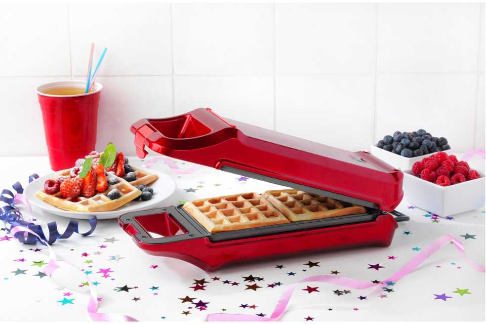 Waffles in a waffle maker near a bowl of blueberries and strawberries on a counter.