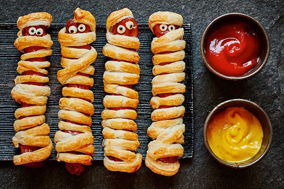 Four sausage rolls with twisted pastry and ketchup and mustard dips.