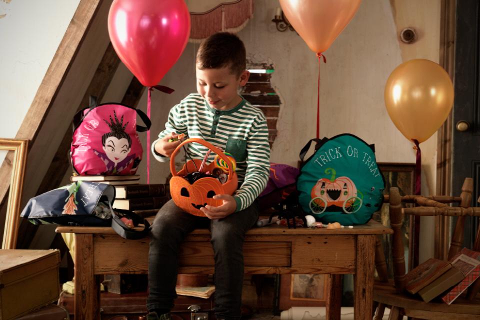 A child sitting on a table picking out sweets from his pumpkin basket. Halloween bags and balloons surround him.