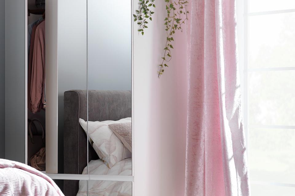 White wardrobe with mirrored door panels next to window with pink curtains.
