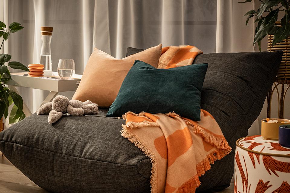 Image of a grey bean bag chair with a couple of cushions an orange throw.