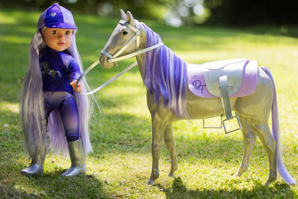 18 inch DesignAfreind doll in a purple horse riding outfit in a field, holding the reins of her DesignAfriend horse with purple mane. 