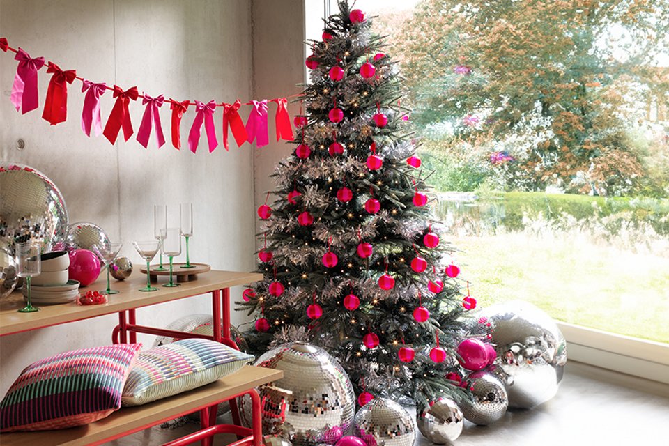 A Christmas tree next to a window decorated in pink and silver with shiny disco balls underneath.