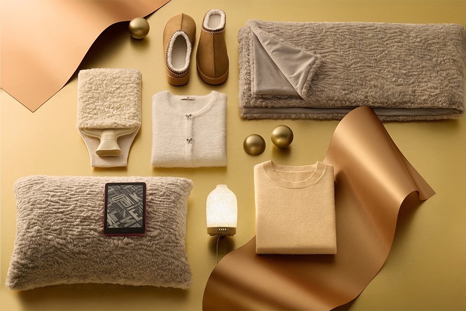 Flat lay of cozy winter essentials in neutral tones, including fluffy beige pillow with e-reader, soft throw blanket, cream cardigan, folded sweater, warm slippers, and decorative gold accents on a golden background.