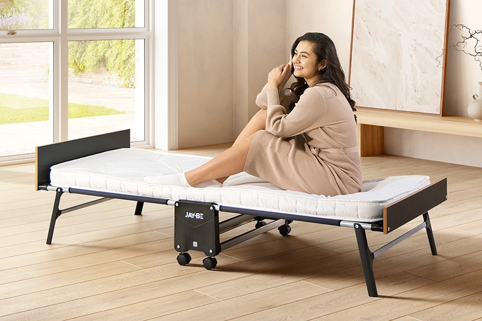 A woman sitting on Jay-Be® Grand folding single bed with e-Pocket™ mattress in a bedroom setup. 