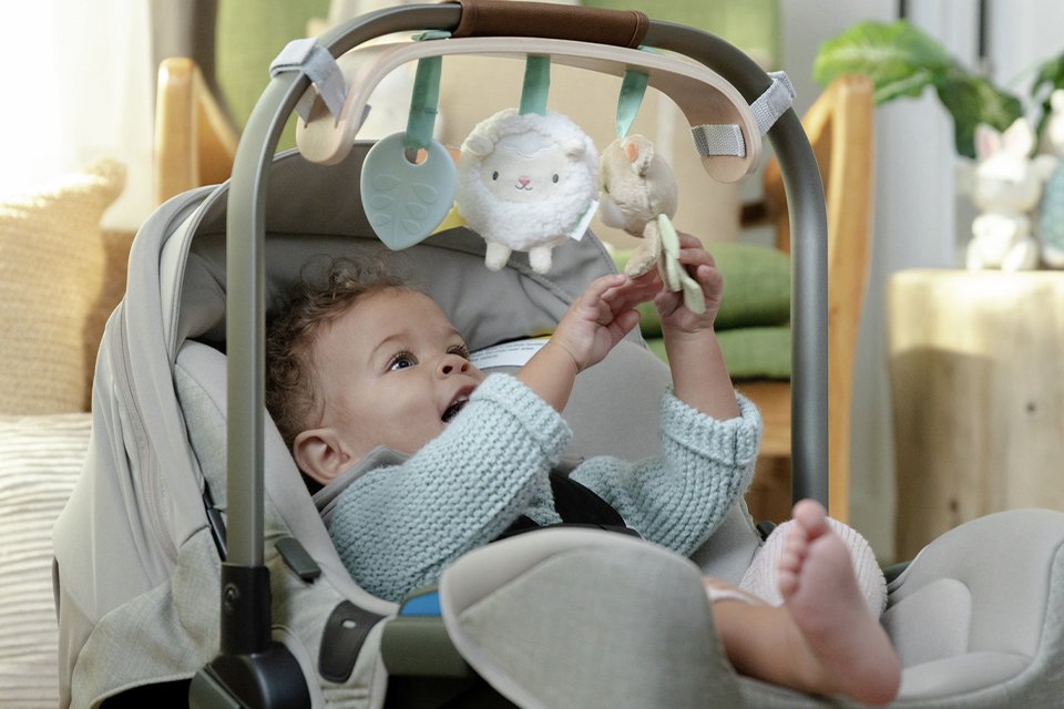 A baby playing with an Ingenuity Wooden Toy Bar attached to a car seat.