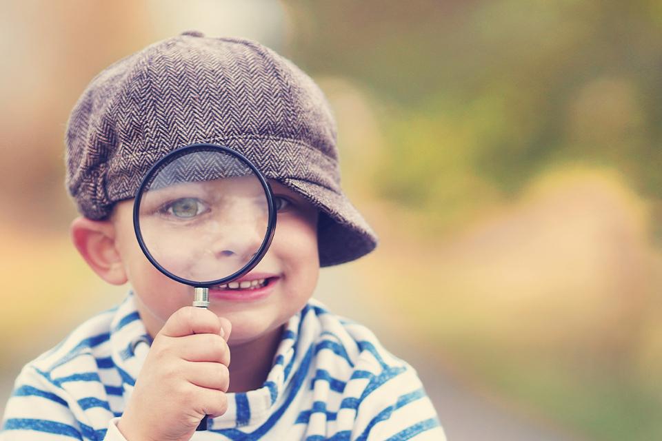 Young boy in blue & white striped top, wearing a grey hat with the peak to the side, looking through magnifying glass at the camera. 