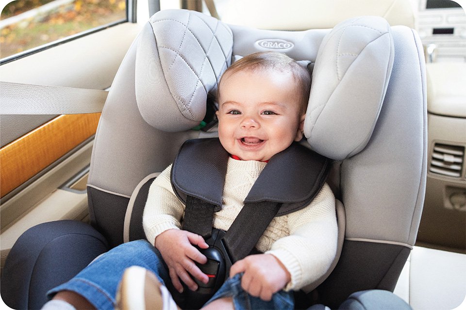 A smiling baby sitting on a Graco SlimFit R129 car seat.