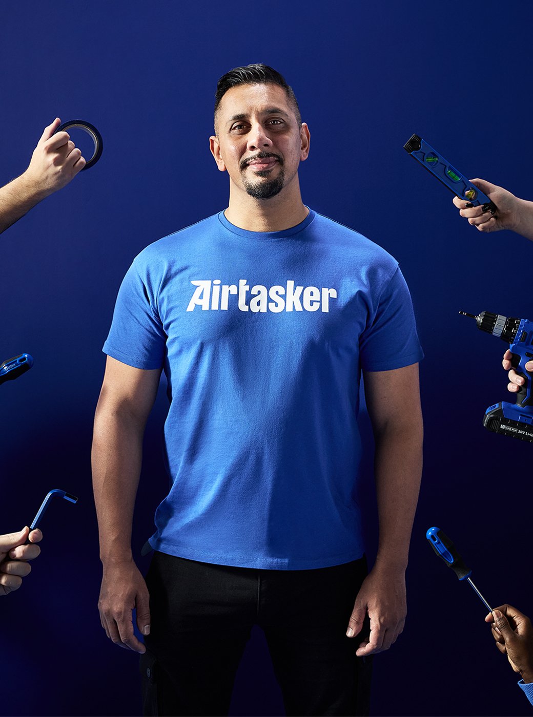Air tasker man stood in a blue room with hands holding building tools out towards him from every angle.