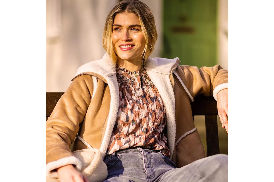 Person sitting on a wooden bench wearing a tan shearling-lined jacket over a patterned blouse, paired with high-waisted light blue jeans and holding a cream textured bag.