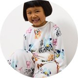 Child wearing white pyjamas featuring colourful cartoon dog prints, sitting on a light surface with pink slippers.