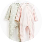 Two baby sleepsuits hanging on white hangers, one in pastel green with white heart pattern and the other in pink and white stripes, both featuring zip-up fronts and footed design.