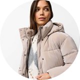 Person wearing a beige puffer jacket with a hood, layered over a white and gray patterned knit sweater, paired with dark brown trousers. 