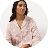 Person wearing a light pink button-up pyjama set with an all-over floral print, sitting against a plain white background.