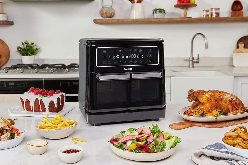 Breville air fryer placed on white kitchen counter with plates of food surrounding it.