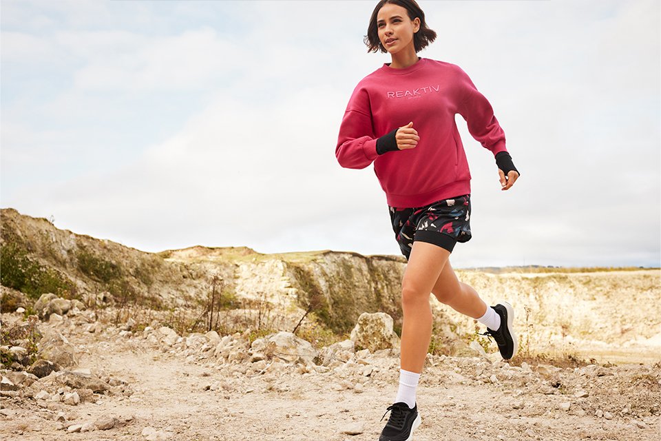 Person running outdoors on a rocky trail, wearing a bright pink sweatshirt with "REAKTIV" text, patterned shorts, white socks, and black running shoes.