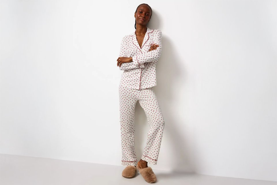  Person standing against a white wall wearing a white pajama set with small black and red heart prints, red piping detail, and beige fluffy slippers. 