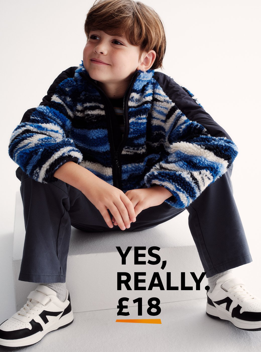 Person sitting on a white block wearing a blue, black, and white patterned zip-up jacket, dark pants, and white sneakers with black accents.