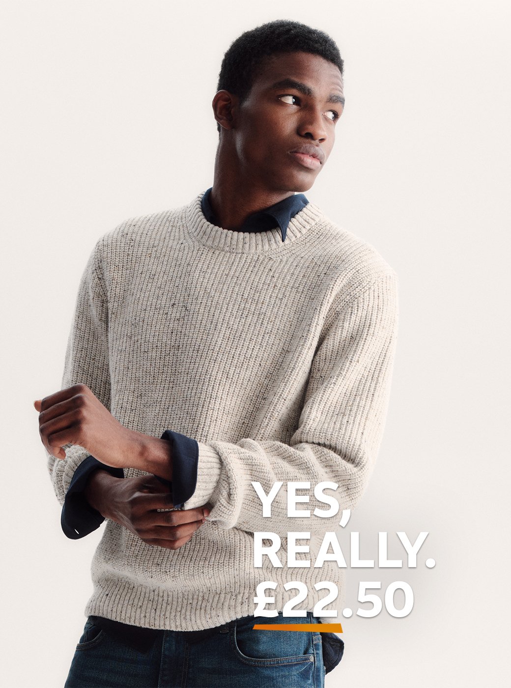 A man wearing a neutral jumper and a dark shirt, with jeans. 