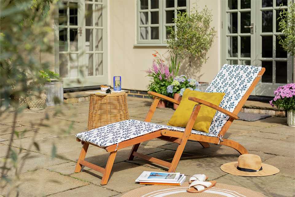An Argos Home folding white wooden sun lounger with a yellow pillow on it in a garden.