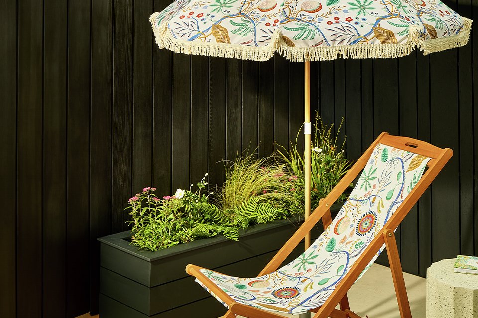 Scion x Habitat garden umbrella and deck chair in white floral pattern next to garden planter with green plants.