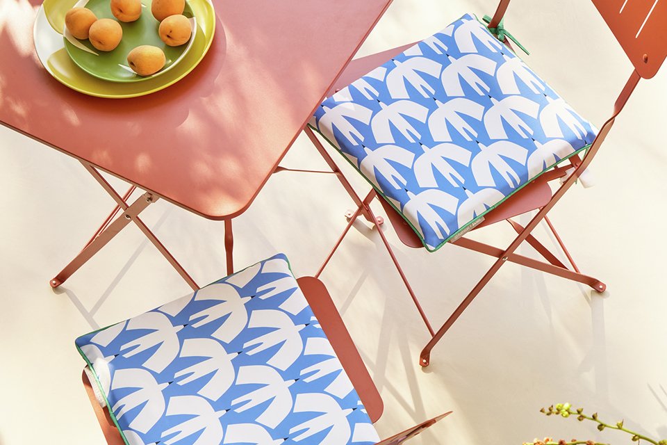 Two oragen harden seats with blie bird patterned cushion next to an orange outdoor table creating a cosy outdoor dining space perfect for breakfast in the sun as part of the Scion x Habitat collection.