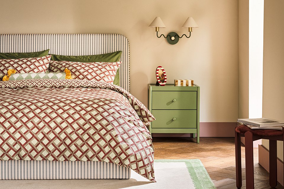 Neutral bedroom with geometric patterned bedding with striped headboard as a vocal point and sage green bed side table with two arm wall light with white shades.