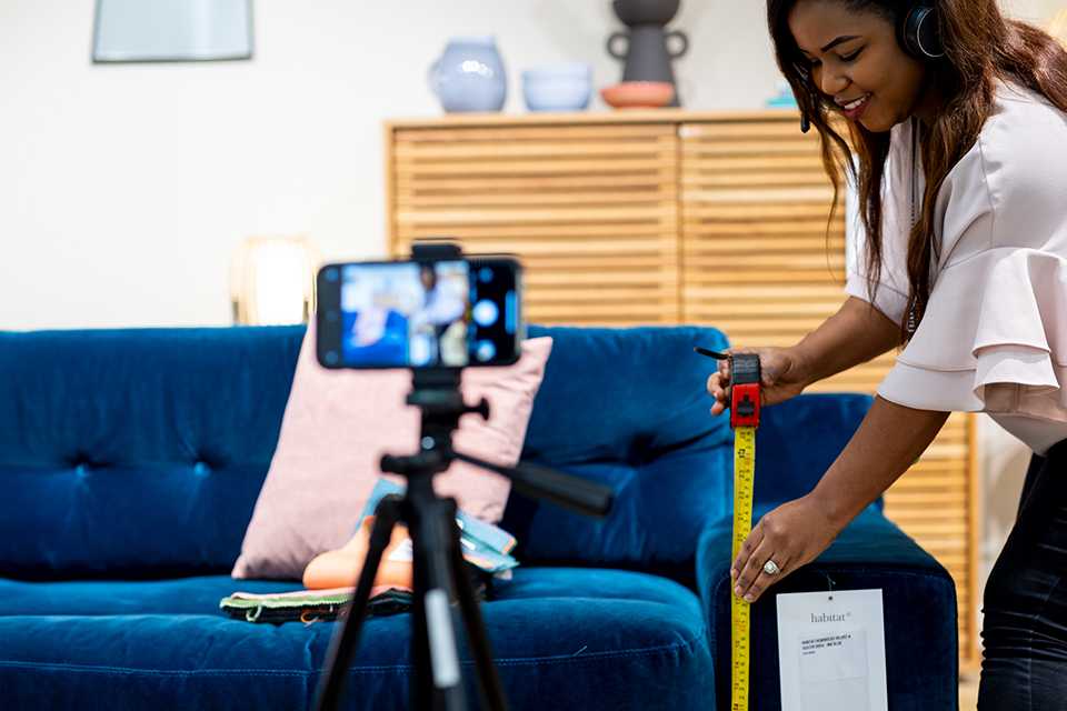 A woman measuring a sofa while being recorded on a phone.