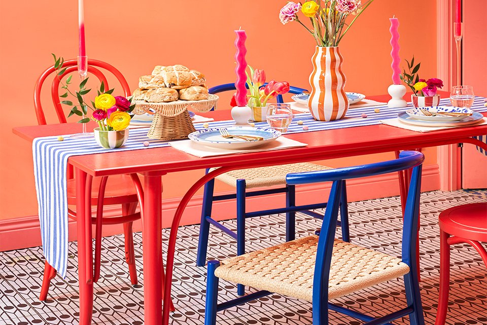 A red colour dining table decorated for Easter with candles, vase, a cake stand, dinnerware, and a table runner. 
