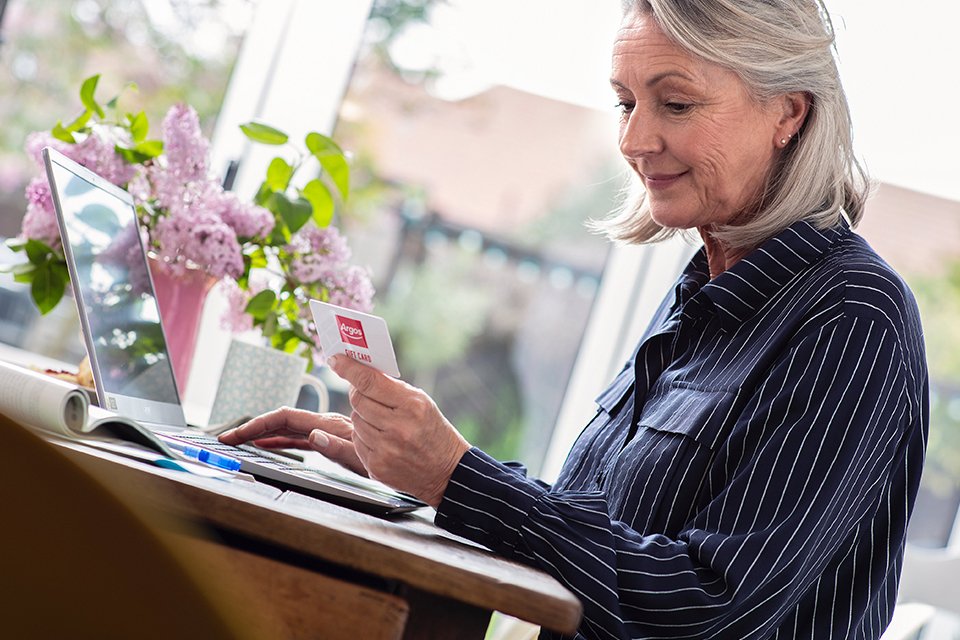 Woman holds argos gift card whilst using a laptop.
