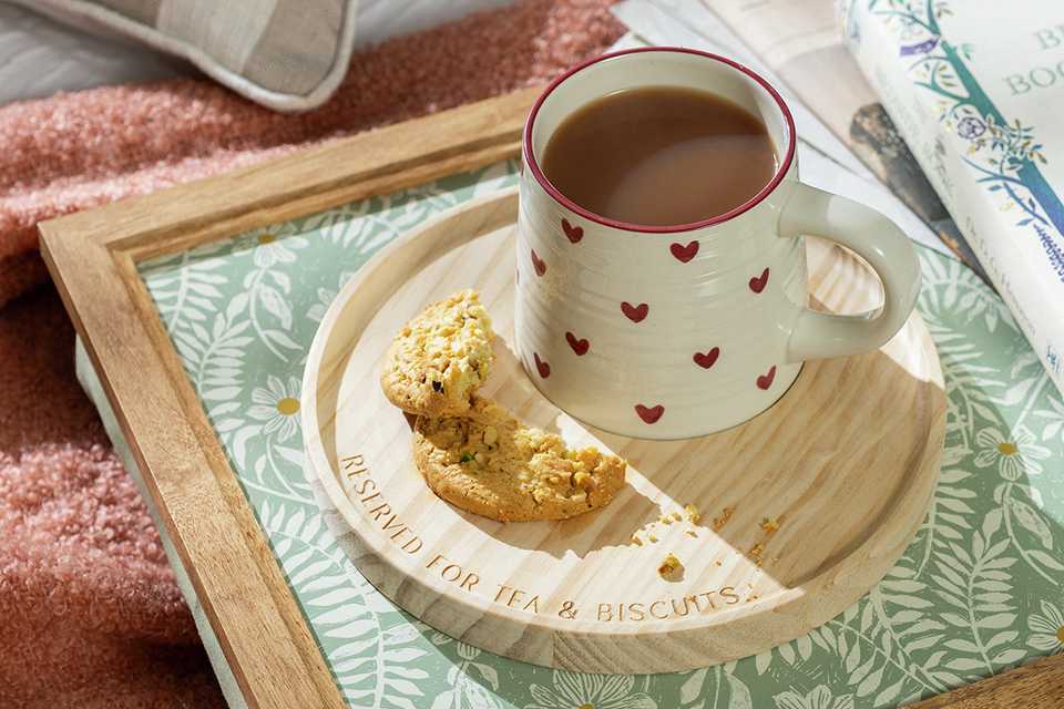 A Home Wooden Tea & Biscuits Tray on a bed with a cookie and mug on top of it.