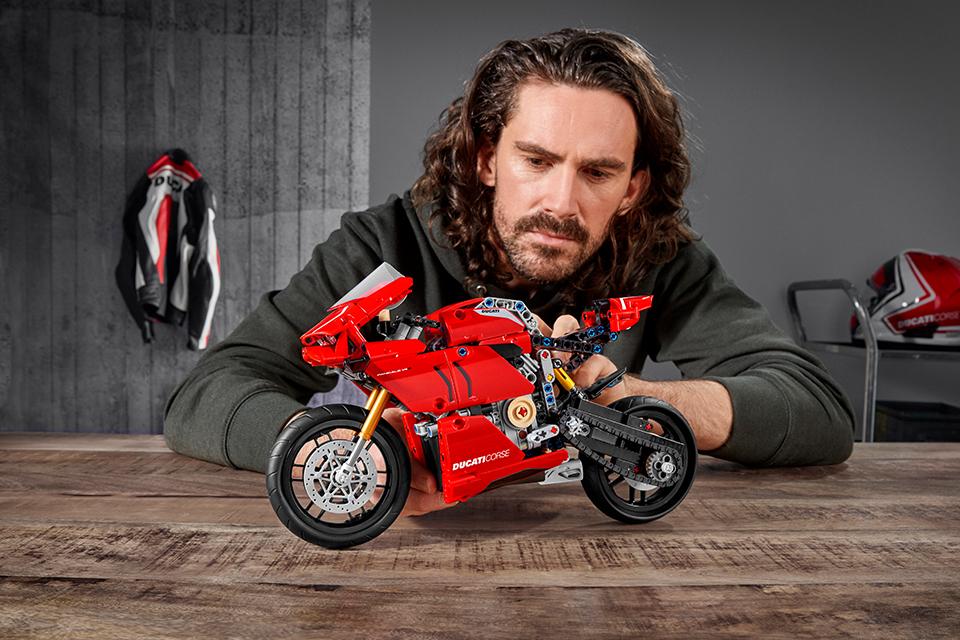 A man with long hair sits at a wooden table. He's holding a red LEGO® Technic model of the Ducati Panigale motorbike.