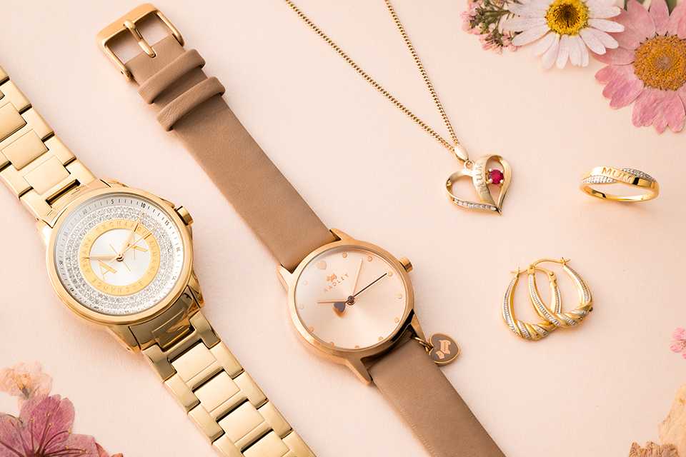A gold watch, rose gold watch and some jewellery on a pink background with flowers.