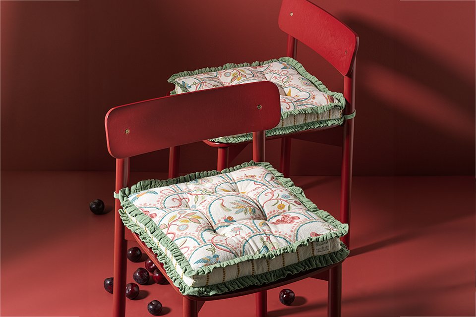 Red chair with white floral printed seat cushion with green frill edge as part of a 2 pack with the chair placed in a red room.
