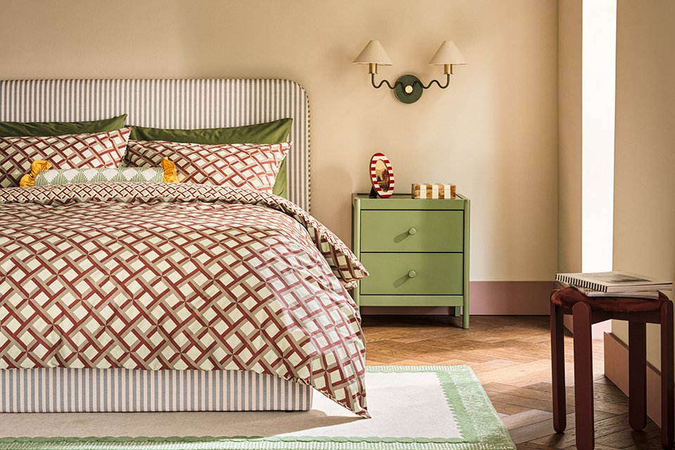 Neutral bedroom with geometric patterned bedding with striped headboard as a vocal point and sage green bed side table with two arm wall light with white shades.
