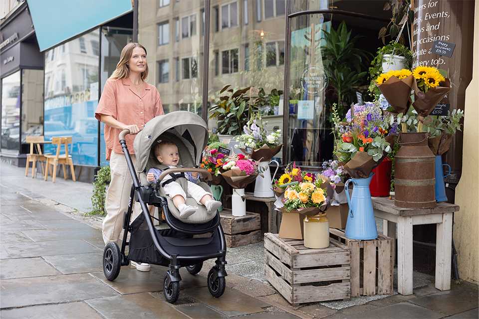 A smiling baby in a Joie versatrax™ pushchair being pushed by mum past a florist shop.