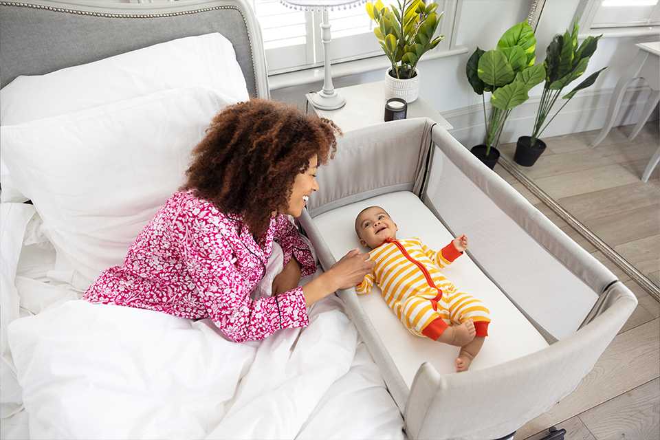 A mother in bed checking on happy baby in a roomie™ go travel bedside crib.